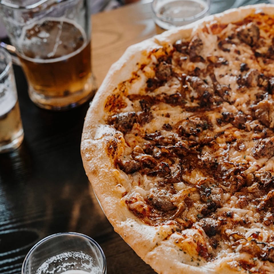 A large, hot pizza with meat and cheese, accompanied by several glasses of beer on a dark table.